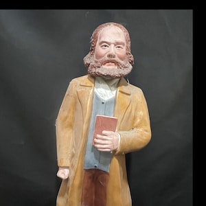 May include: A painted figurine of a bearded individual with a receding hairline, wearing a tan overcoat, a grey waistcoat, and a white collared shirt. The figure holds a small, rectangular red book in one hand and has a serious facial expression.