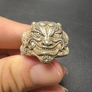 Op de afbeelding: Een zilverkleurige ring met het gedetailleerde, gebeeldhouwde gezicht van een mythisch wezen met opvallende hoektanden, grote ogen en wervelende patronen. Het metalen oppervlak is gepolijst met donkere verdiepte delen om het ontwerp te accentueren.