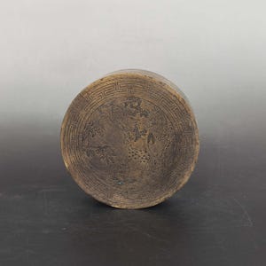Könnte beinhalten: Ein runder, bronzefarbener Metallbehälter mit einem gravierten Deckel. Der Deckel zeigt eine zentrale Szene mit einem gefleckten Hirsch zwischen Bäumen, umgeben von einem geometrischen Mäander-Muster, das in die dunkle Metalloberfläche geätzt ist.