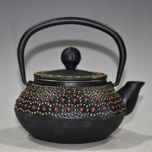 May include: A black cast iron teapot featuring a rounded body and a matching lid with a spherical knob. The exterior surface is decorated with a repeating pattern of small, raised red dots surrounded by yellow painted accents on the lid and body.