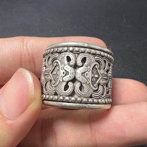 Può includere: Un ampio anello in metallo color argento caratterizzato da un intricato motivo simmetrico in rilievo di ali o volute stilizzate. La fascia è bordata da file di piccole perle sferiche in rilievo sui bordi superiore e inferiore, con una finitura ossidata scura.