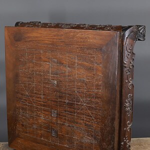 May include: A dark brown wooden Chinese chess board featuring a grid pattern with engraved lines and Chinese characters. The board has a thick, carved frame with intricate scrollwork and decorative patterns along the side edges and top border.