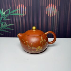May include: A round, reddish-brown clay teapot featuring a gold-coloured knob on the lid and a curved handle. The body of the teapot is decorated with a delicate, hand-painted gold floral and bamboo design that wraps around the front surface.