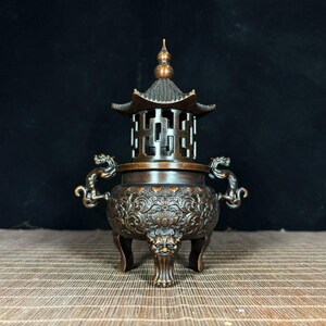 Puede incluir: Un quemador de incienso de bronce oscuro con una intrincada tapa estilo pagoda con un remate, dos asas laterales en forma de dragón y un cuerpo redondo decorado con relieves florales detallados, sostenido por tres patas con forma de león.