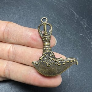 May include: A bronze-toned metal pendant shaped like a ritual axe, featuring an ornate handle with a loop at the top and a curved, textured blade decorated with intricate swirling patterns and embossed details across the entire surface.