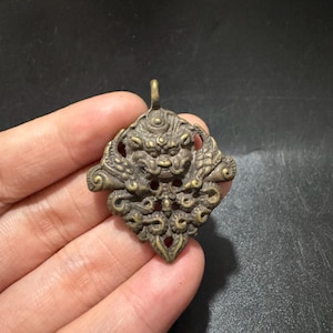 Può includere: Un ciondolo in metallo color bronzo con un rilievo dettagliato di una creatura mitica dal volto prominente e motivi ornamentali a spirale. Il ciondolo presenta un piccolo occhiello sulla parte superiore e una finitura con patina scura.