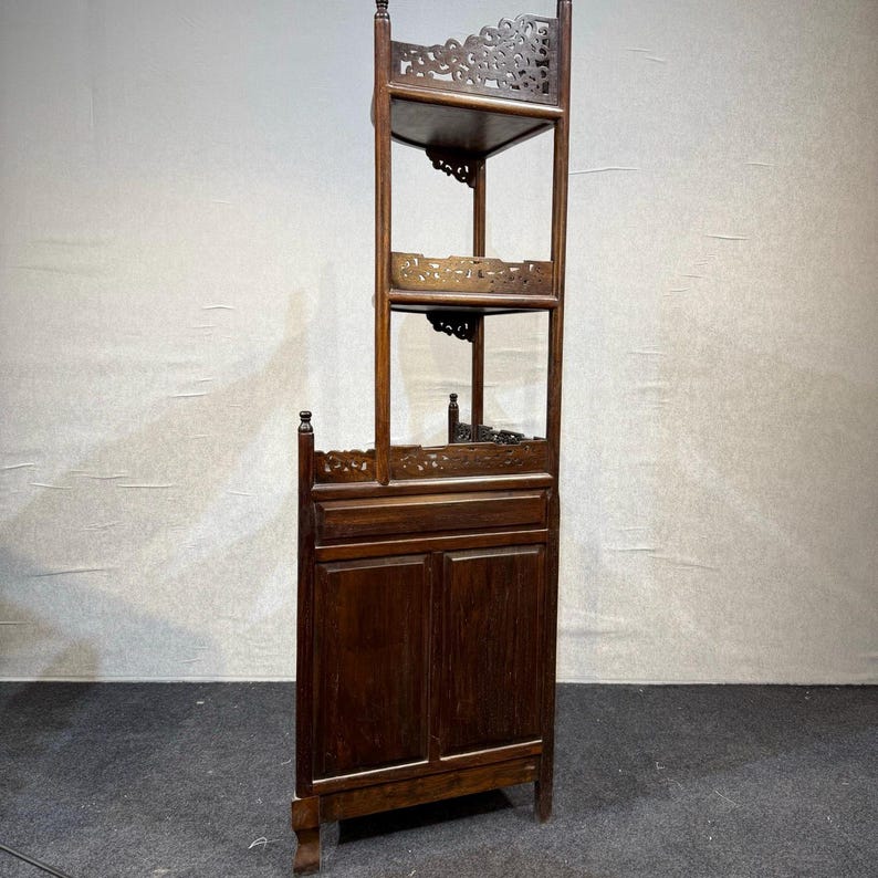May include: A tall, dark brown wooden corner cabinet featuring a lower section with two solid doors and an upper section with two open shelves. The unit includes intricate carved lattice detailing along the top edges of the shelves and the base.