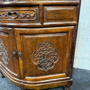 May include: A dark brown wooden corner cabinet featuring a curved front, a single drawer with an ornate carved handle, and two doors decorated with intricate, symmetrical scrollwork patterns. The cabinet has a polished wood grain finish.