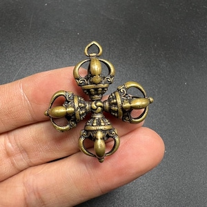 May include: A brass-coloured metal pendant shaped like a four-pronged vajra or dorje. The object features a central hub with four symmetrical, curved arms extending outwards, each ending in a pointed tip. It has a small loop at the top for hanging.
