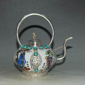 May include: A white ceramic teapot with a silver metal handle, spout, and base. The body features colorful painted figures, black Chinese calligraphy, and inlaid turquoise stones. A silver metal filigree ornament with a central turquoise stone is attached.