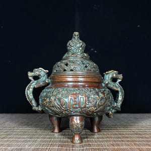 May include: A bronze-toned metal incense burner featuring a lid topped with a sculpted figure, two dragon-shaped side handles, and three legs. The body is decorated with intricate relief carvings of dragons and a central rectangular Chinese character.