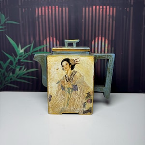 May include: A square-shaped teapot with a weathered blue-green finish and gold-toned edges. The front panel features a detailed illustration of a person with dark hair styled with ornaments, wearing a flowing robe, set against a light background.