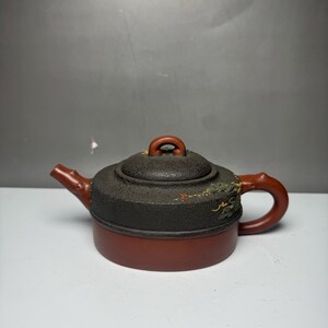 May include: A two-toned teapot featuring a smooth, reddish-brown cylindrical base and a textured, dark charcoal-colored lid. The teapot includes a branch-shaped handle and spout, with small, delicate gold and green painted floral accents on the lid.