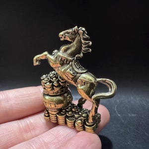 May include: A small brass-coloured metal figurine depicting a horse rearing up on its hind legs. The horse is positioned atop a stack of circular coins and a rounded container. The metal has a weathered, antique finish with dark highlights.