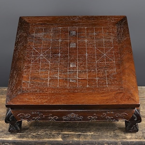 May include: A dark brown wooden Xiangqi board featuring a grid of thin white lines and Chinese characters etched into the surface. The board has a raised border and is supported by four carved legs with decorative scrollwork along the apron.
