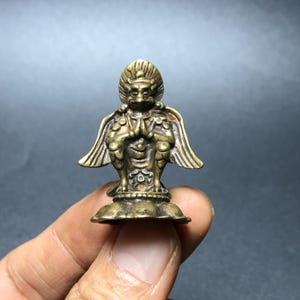 Può includere: Piccola e dettagliata statuetta in ottone di una creatura mitica con volto di uccello, ali e mani giunte in preghiera. La figura è montata su una base a forma di fiore di loto, che mostra dettagli intricati.