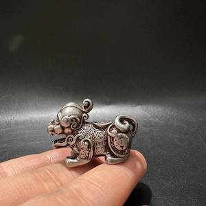 Op de afbeelding: Een klein, zilverkleurig metalen beeldje van een mythisch wezen, mogelijk een leeuw of draak, met ingewikkelde reliëfpatronen, een gekrulde staart en een gedetailleerd gezicht met een open mond, in een compacte, sculpturale vorm.