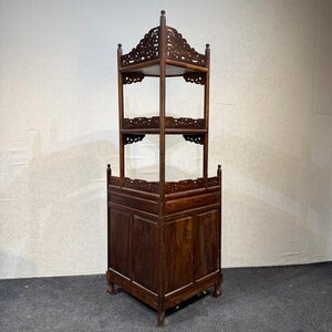 May include: A tall, dark brown wooden corner cabinet featuring a solid base with paneled doors and two open upper shelves. The structure includes intricate carved fretwork along the top edges of each shelf and the base, supported by four legs.
