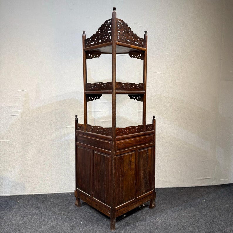 May include: A tall, dark brown wooden corner cabinet featuring a solid base with paneled doors and two open upper shelves. The structure includes intricate carved fretwork along the top edges of each shelf and the base, supported by four legs.