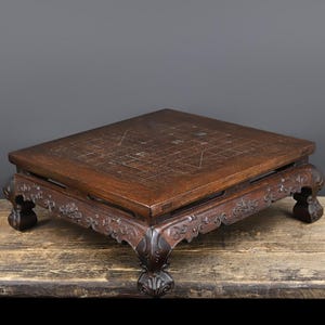 May include: A square wooden game board featuring a grid pattern etched into the surface. The board is supported by four ornate, carved legs with scrollwork details along the apron. The dark brown wood has a visible grain and polished finish.