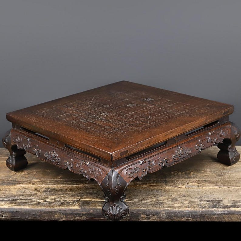 May include: A square wooden game board featuring a grid pattern etched into the surface. The board is supported by four ornate, carved legs with scrollwork details along the apron. The dark brown wood has a visible grain and polished finish.