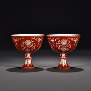 Puede incluir: Un par de copas de tallo rojo intenso con patrones florales y de enredaderas en blanco. Cada copa tiene un cuenco ancho y acampanado y un tallo alto y esbelto decorado con motivos de hojas blancas y pequeños detalles florales.