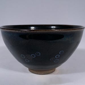 May include: A deep, rounded ceramic bowl with a glossy black glaze. The exterior features small, faint blue circular patterns arranged in clusters. The rim is dark brown, and the base is an unglazed, light brown circular foot that elevates the bowl.
