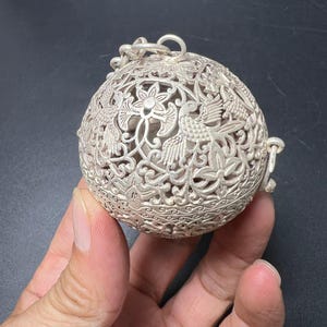 May include: A spherical silver-toned metal pendant featuring intricate openwork filigree patterns. The design displays detailed carvings of birds and floral motifs across the surface. A small metal loop is attached to the top for hanging.