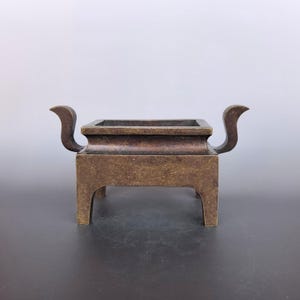 Pode incluir: Um queimador de incenso retangular de bronze com um acabamento met&aacute;lico escuro e desgastado. O recipiente tem uma borda larga e plana, quatro pernas robustas e duas al&ccedil;as curvas proeminentes que se estendem para os lados.