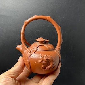 May include: A terracotta-colored clay teapot featuring a high, arched handle with a textured, branch-like appearance. The body of the teapot is adorned with a relief carving of a dragon, and the lid is topped with a small, mushroom-shaped finial.