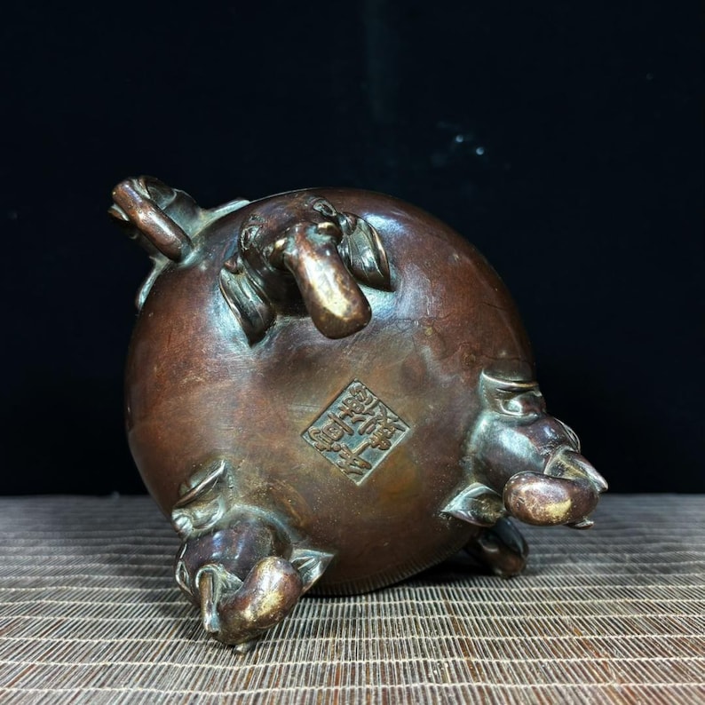 K&ouml;nnte beinhalten: Ein runder R&auml;uchergef&auml;&szlig; aus Bronze mit drei elefantenf&ouml;rmigen Beinen und einem quadratischen Siegelstempel auf der Unterseite. Die Metalloberfl&auml;che weist eine dunkelbraune Patina mit helleren metallischen Akzenten an den Elefantenr&uuml;sseln auf.