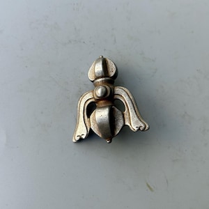 May include: A silver-colored, ornate metal object with a symmetrical design. It features a central, rounded body with stylized wings or curved elements extending outwards. The top has a rounded, crown-like shape.