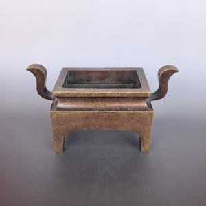 Pode incluir: Um queimador de incenso retangular de bronze com um acabamento met&aacute;lico escuro e desgastado. O recipiente tem um topo largo e aberto, duas al&ccedil;as curvas que se estendem para os lados e quatro pernas integradas e robustas na base.