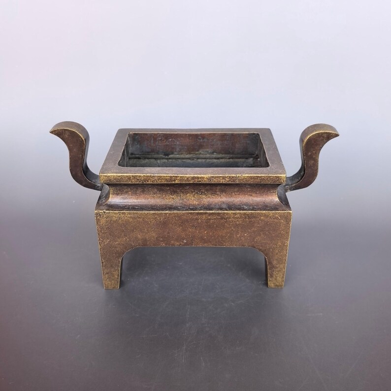 Pode incluir: Um queimador de incenso retangular de bronze com um acabamento met&aacute;lico escuro e desgastado. O recipiente tem um topo largo e aberto, duas al&ccedil;as curvas que se estendem para os lados e quatro pernas integradas e robustas na base.
