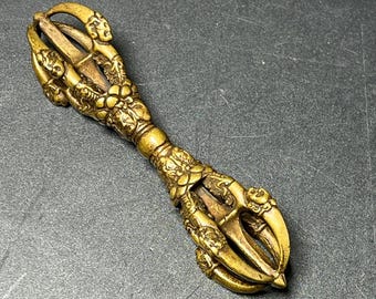 May include: A metallic gold-toned vajra ritual object featuring intricate relief carvings of faces and floral motifs. The symmetrical design consists of a central handle with two identical multi-pronged ends, each showcasing curved prongs meeting at a point.
