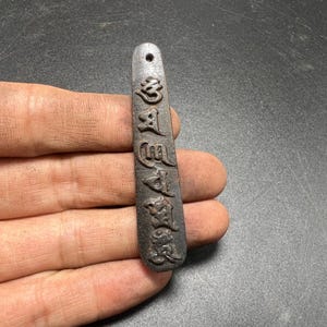 May include: A long, narrow, dark brown metal pendant with a rounded top and a small hole for suspension. The surface features a vertical column of five raised, embossed characters in a script style, creating a textured, tactile appearance.