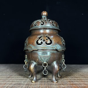 May include: A bronze-toned metal incense burner featuring a removable lid with intricate cut-out patterns. The vessel has a rounded body with embossed decorative motifs, supported by three curved legs, each adorned with a small dangling metal ring.