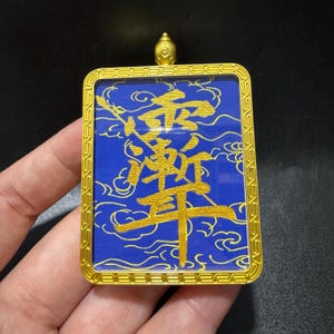 Könnte beinhalten: Ein rechteckiger Anhänger mit einem leuchtend blauen Hintergrund, goldenen chinesischen Schriftzeichen und wirbelnden Wolkenmustern. Das Stück ist in einen polierten, goldfarbenen Metallrahmen mit geometrischer Bordüre und Öse gefasst.