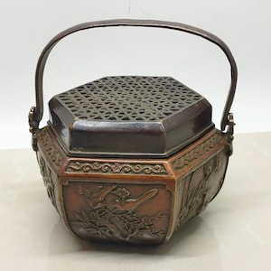 May include: A hexagonal bronze incense burner featuring a dark patina and a swing handle. The body displays relief carvings of birds perched on flowering branches. The lid has an intricate openwork lattice pattern, allowing for smoke release.