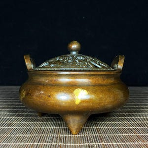 Pode incluir: Um queimador de incenso de metal em tom de bronze com corpo arredondado, três pernas curtas e duas alças laterais. A tampa removível tem um design intrincado e vazado com um botão central. Uma pequena marca amarela brilhante é visível.