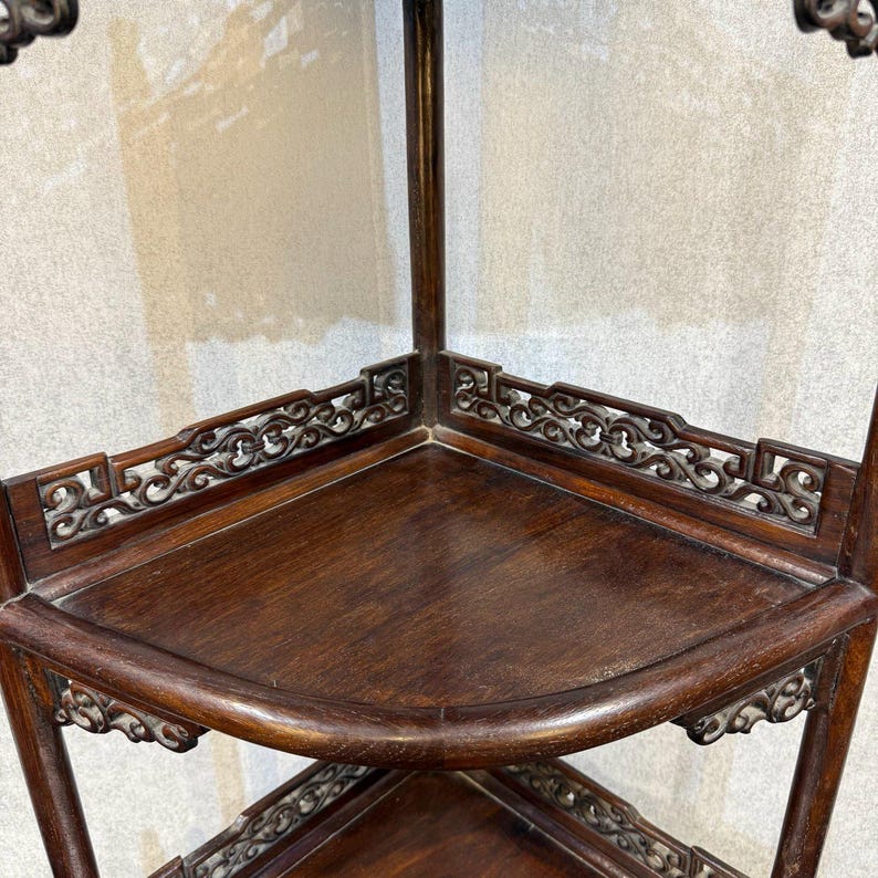 May include: A dark brown wooden corner shelf unit featuring multiple tiers with curved edges. Each shelf is framed by intricate, openwork carved lattice detailing along the back edges and decorative scrollwork supports beneath the shelf surfaces.