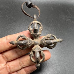 May include: A dark, weathered metal vajra pendant featuring a cross-shaped design with four symmetrical prongs extending from a central hub. The object has a textured, aged surface and is attached to a small, worn black leather cord loop at the top.