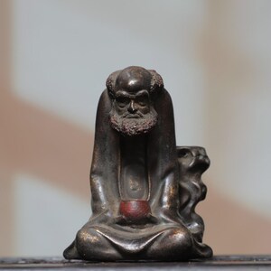 May include: A dark brown bronze-toned figurine of a seated figure with a textured beard and bald head, wearing a draped robe. The figure holds a small, smooth, reddish-brown bowl in both hands, with a stylized animal form positioned to the right.