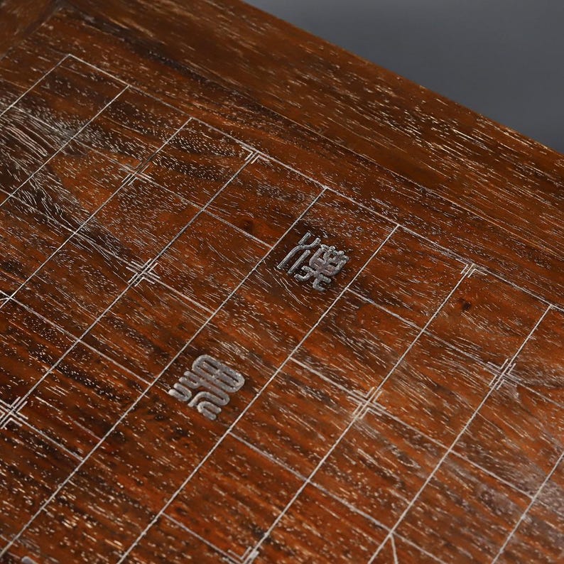 May include: A dark brown wooden board game surface featuring a grid of thin white lines. The board displays two distinct Chinese characters engraved or inlaid in a light metallic color, positioned within the central grid squares of the wood.