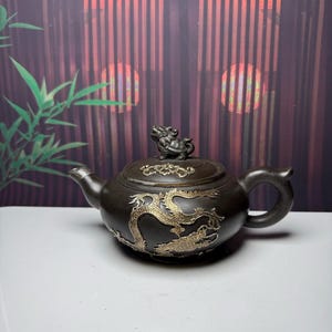Peut inclure: Une théière brun foncé ornée d'un relief de dragon doré sur le corps, dotée d'une anse incurvée, d'un bec court et d'un couvercle surmonté d'une petite figurine de créature sculptée. Le couvercle présente aussi un motif floral doré.