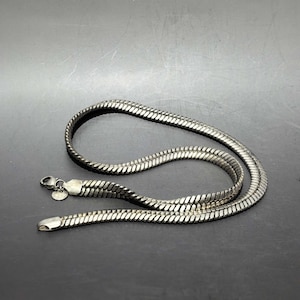 May include: A silver-toned snake chain necklace featuring a flat, flexible design with a polished metallic finish. The necklace includes a secure lobster claw clasp and a small, round metal tag attached near the closure, showing a consistent link pattern.