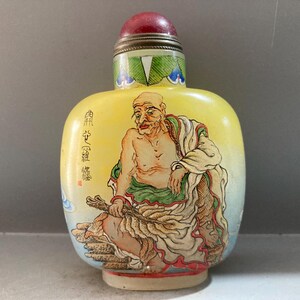 Peut inclure: Une tabatière jaune ornée d'une illustration peinte à la main représentant un personnage assis tenant un éventail. La bouteille a une forme rectangulaire arrondie, un bouchon rouge et des accents décoratifs verts au col. Des caractères chinois sont visibles.
