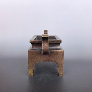 Pode incluir: Um queimador de incenso retangular de bronze com uma bacia profunda, uma borda plana e quatro pernas robustas. A superf&iacute;cie met&aacute;lica apresenta uma p&aacute;tina castanho-escura desgastada com reflexos dourados subtis nas bordas e cantos.