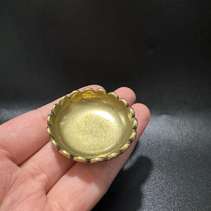 May include: A small, circular, gold-toned metal dish with a scalloped edge. The interior surface of the dish is smooth and reflective, while the outer rim features a series of rounded, petal-like indentations that create a decorative border.