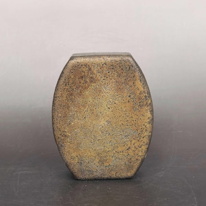 May include: An oval-shaped metal container with a weathered, mottled bronze and brown patina. The surface features a textured, speckled appearance with faint, intricate engraved line patterns visible across the front face of the object.